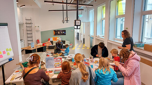 Kinder und Erwachsene sitzen an einem großen Tisch in der Wissenswerkstadt Bielefeld und basteln gemeinsam mit verschiedenen Materialien.