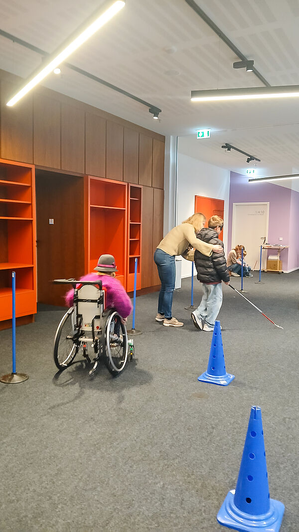 Zwei Kinder durchlaufen einen Parcours, eines im Rollstuhl und eines mit Blindenstock, begleitet von einer erwachsenen Person.