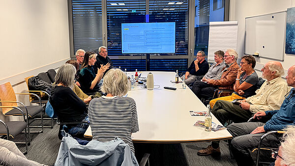 Annemarie Witschas sitzt mit einer Gruppe von Teilnehmenden an einem großen Tisch und diskutiert mit ihnen.
