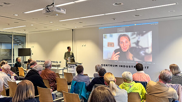 Dr. Theresa Züger ist per Videokonferenz auf der großen Leinwand zu sehen; im Raum sitzt das Publikum vor der Projektion.