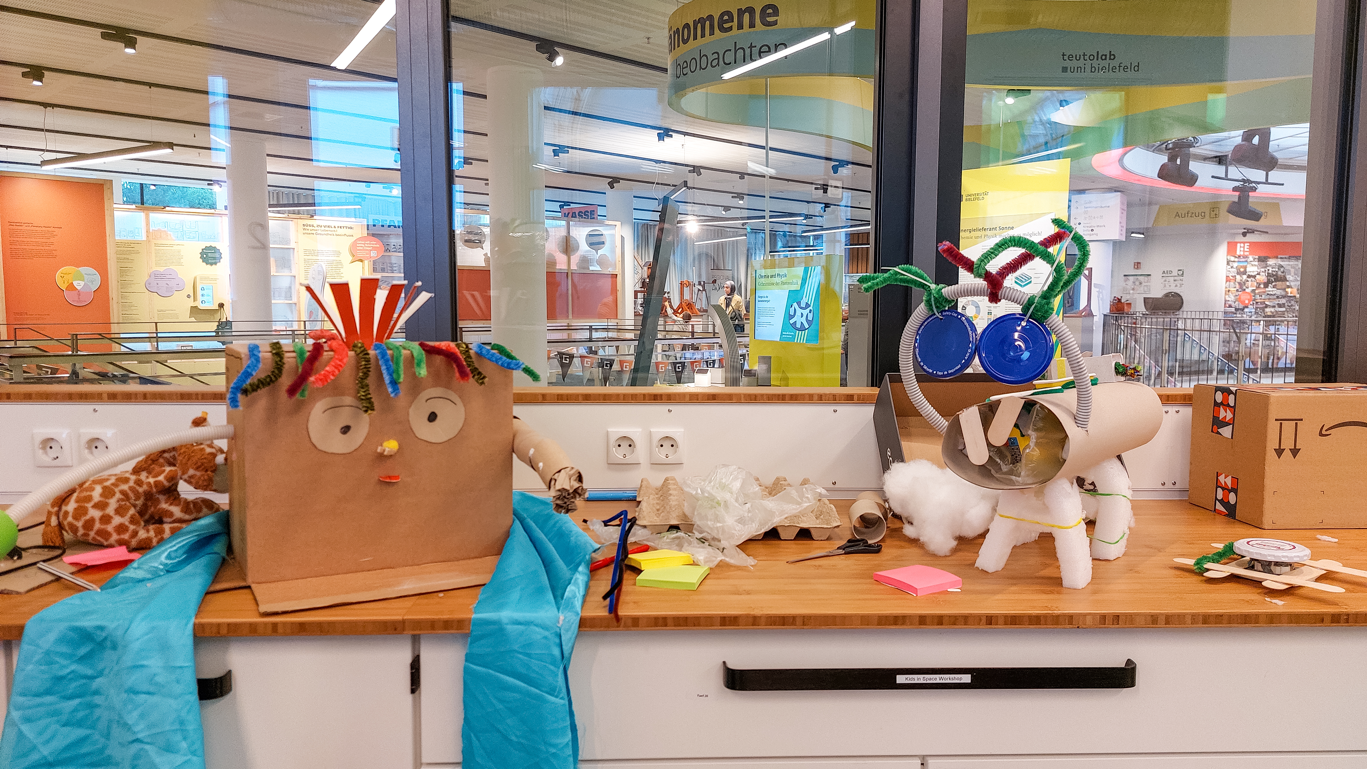 Zwei selbst gebastelte Figuren aus Karton, Papier, Pfeifenreinigern und weiteren Bastelmaterialien stehen auf einem Tisch in der Wissenswerkstadt Bielefeld.