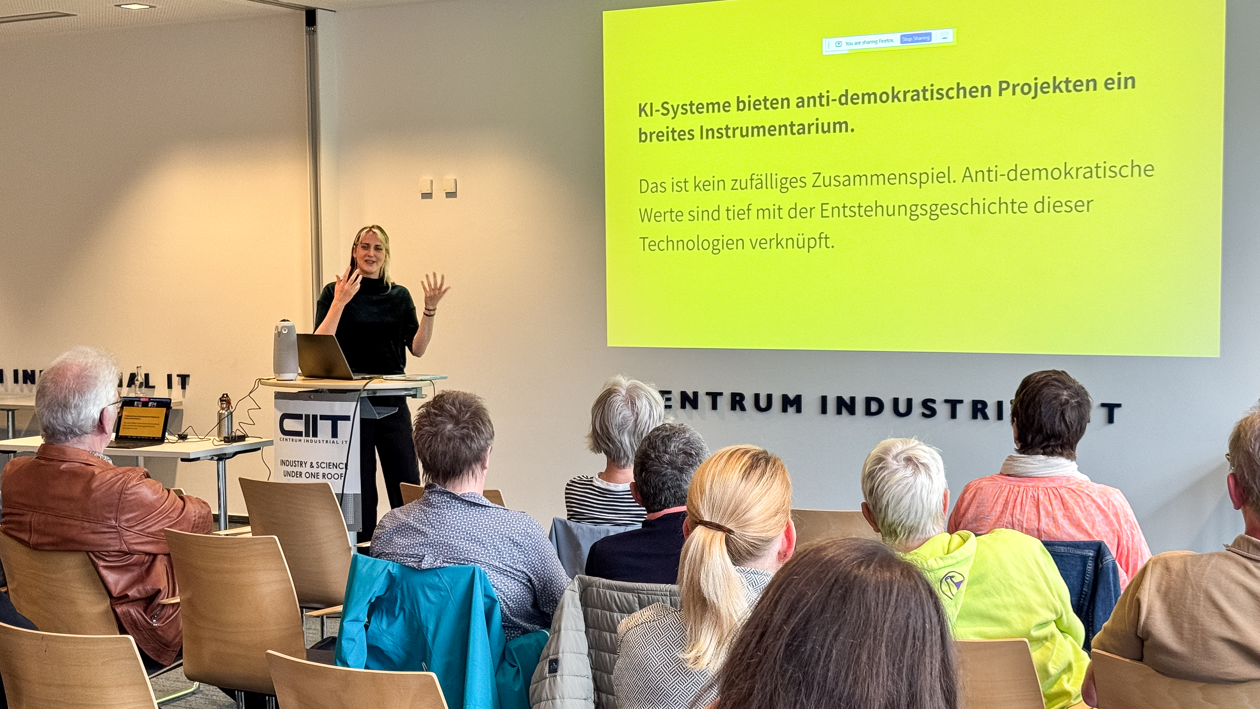 Annemarie Witschas steht vor dem Publikum und spricht neben einer Präsentationsfolie über anti-demokratische Projekte und KI-Systeme.